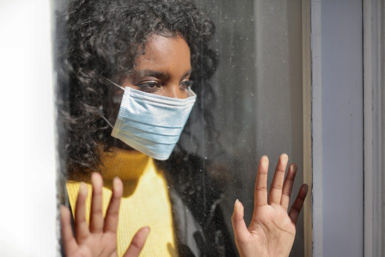 woman looking outside the window wearing facemask due to lockdown